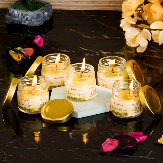 Five small candles in glass jars with gold lids on a dark surface, surrounded by decorative elements.